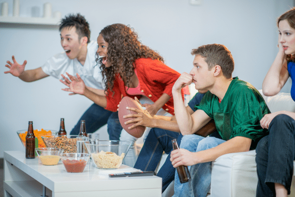 Group of friends watching the Super Bowl very intensely. They are holding a fooball and enjoying some snacks with their attention on the TV.